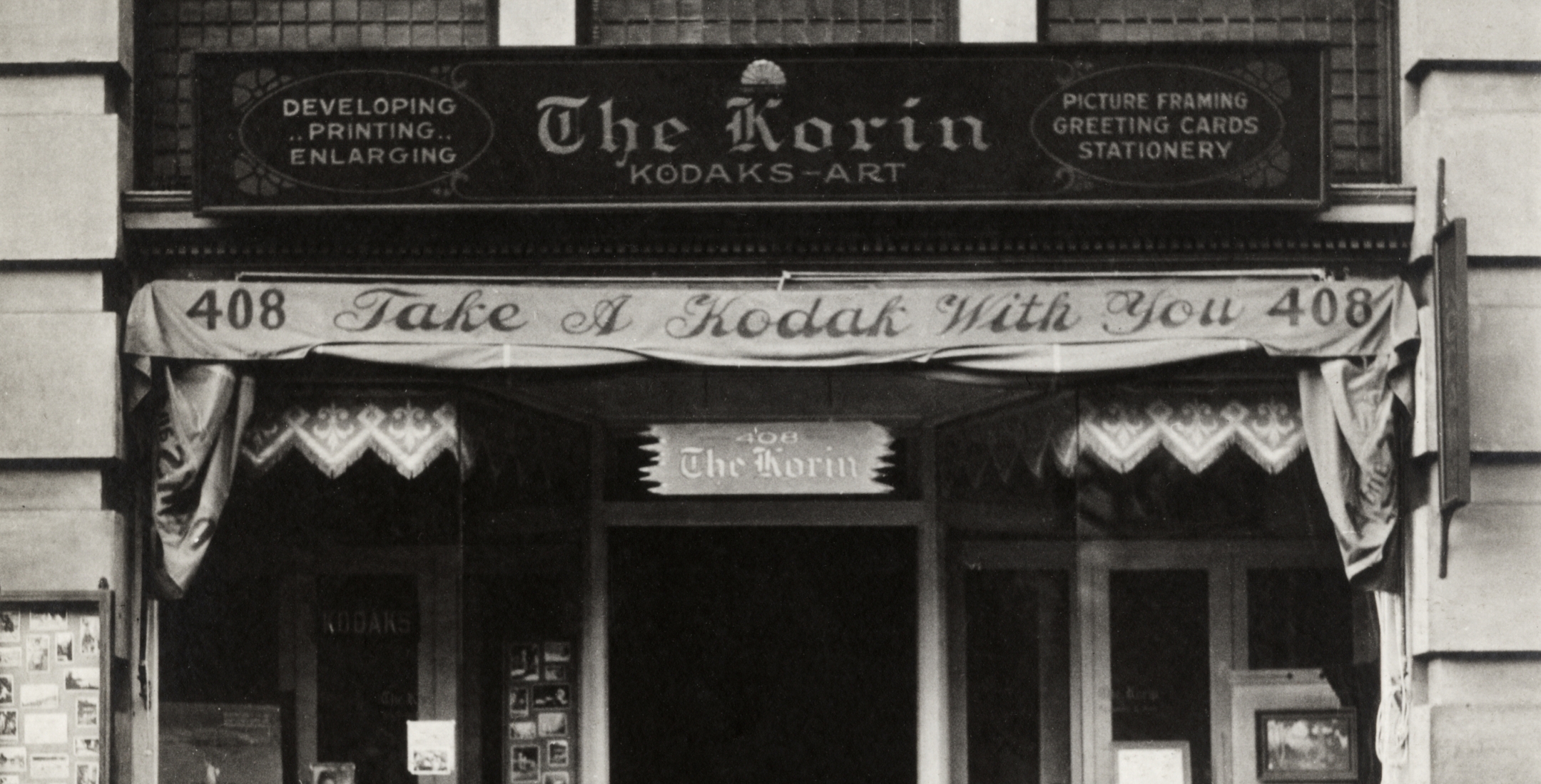 Black and white image of an early 20th century storefront. Signs read: "The Koran KODAKS—ART" and "Take a Kodak with You".