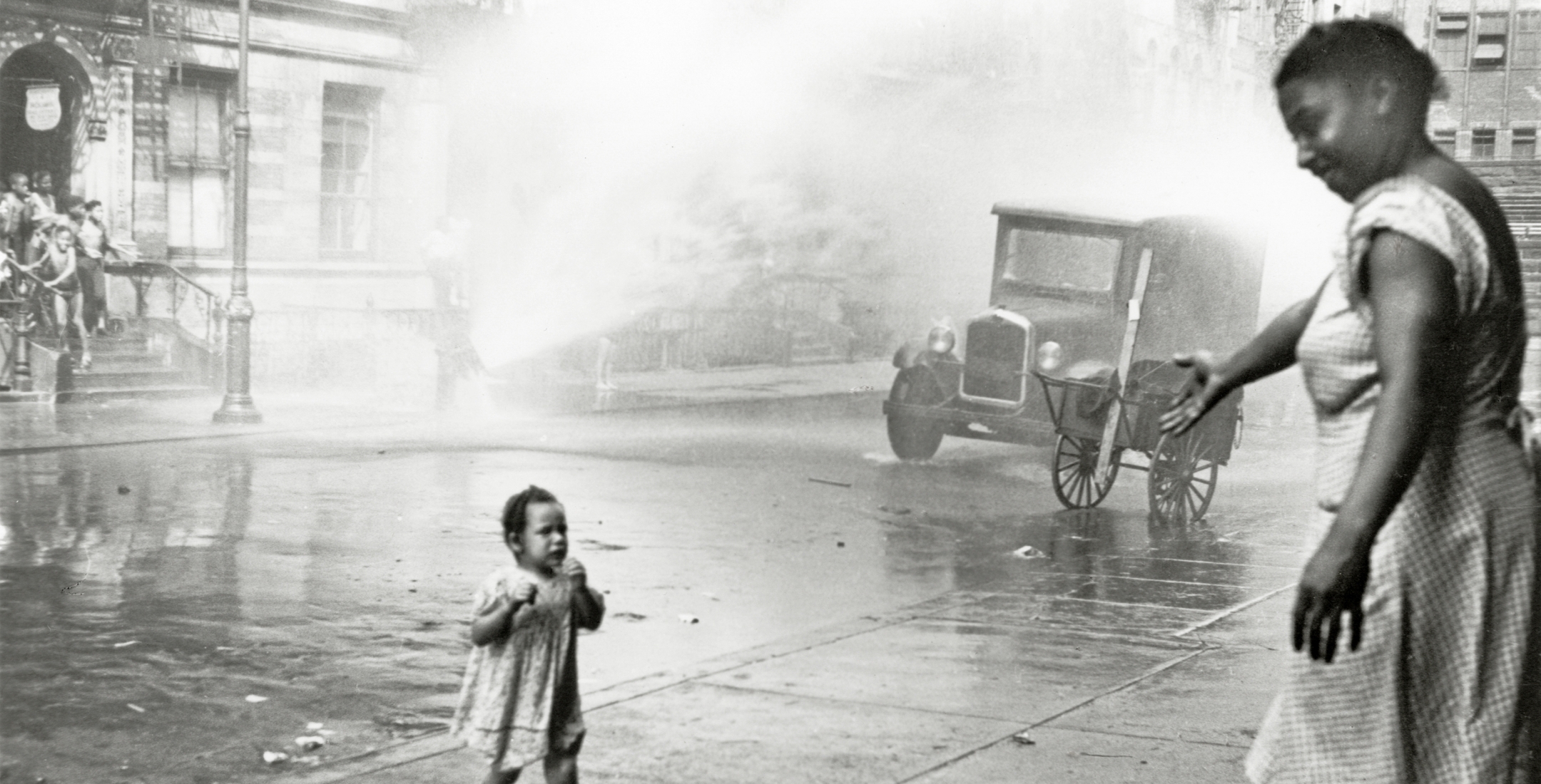Black and white photo of a toddler crossing the street to her mother who waits with outstretched hand, behind her is an open fire hydrant where neighborhood kids are playing.