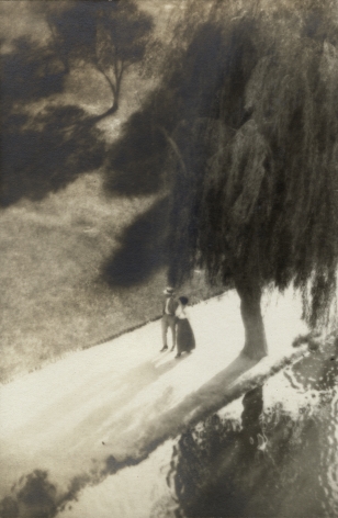 Black and white photo: Two people are seen from above and at a distance, wearing early 20th century weekend clothes and walking around a pond, under a weeping willow tree.