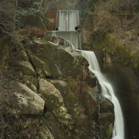 Color photograph of water spillage cascading from a dam in a wooded area.