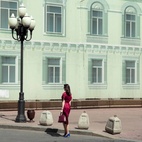Woman in a red dress and blue heels, on a Moscow street, standing with her back to the camera in front of a life-size printed facade of a building.