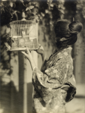 Black and white profile image of a Japanese American woman wearing a Kimono and looking inside a birdcage that she is holding aloft.