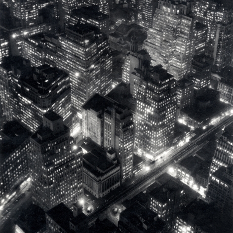Black nd white photo: view looking down from the Empire State building in 1932, an elevated train line runs between lit up buildings at night.