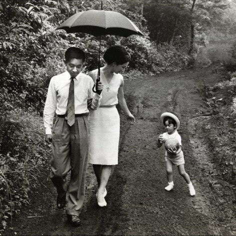 Black and white image of then Crown Prince Akihito of Japan walking on a forest trail and holding an umbrella for his wife as she looks at their son who is holding a toy ball.