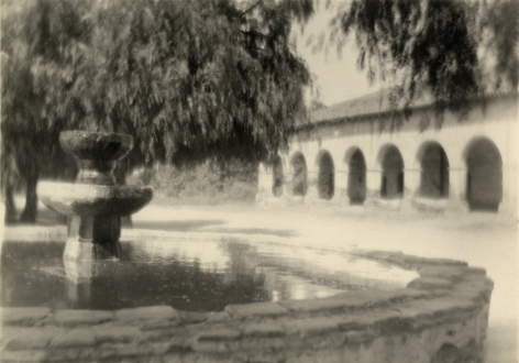 Soft focus black and white photo of a fountain and a pavilion in a park setting with a large weeping willow tree.
