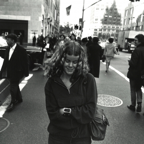 black and white photo shot by Garry Winogrand in 1969 showing a woman smiling and looking down while crossing the street in NYC.
