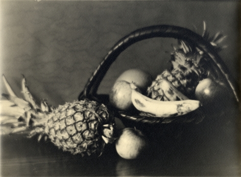 Black and white still life of a handled wicker basket of fruit—pineapple, banana, etc