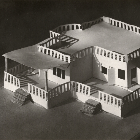 Black and white photo of small architectural model made out of paper–depicting a single story building with a fenced wrap around porch in front.